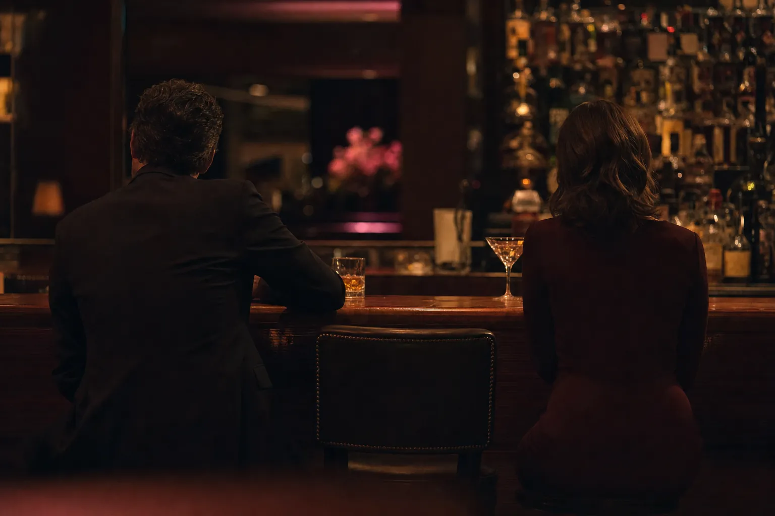 Two people at a dim hotel bar, seen from behind, with an empty stool between them